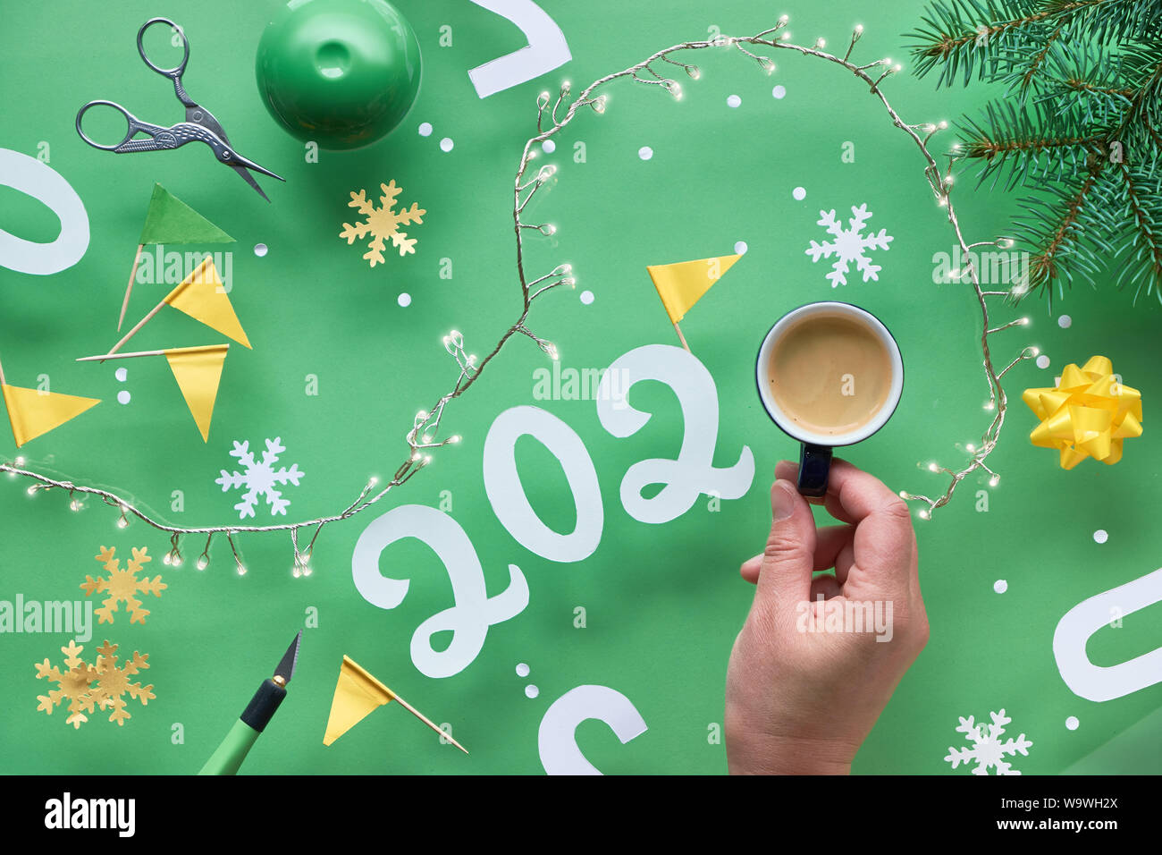 Green and yellow creative Christmas flat lay with number 2020, cup of coffee, hand, Xmas lights and paper craft elements. Stock Photo