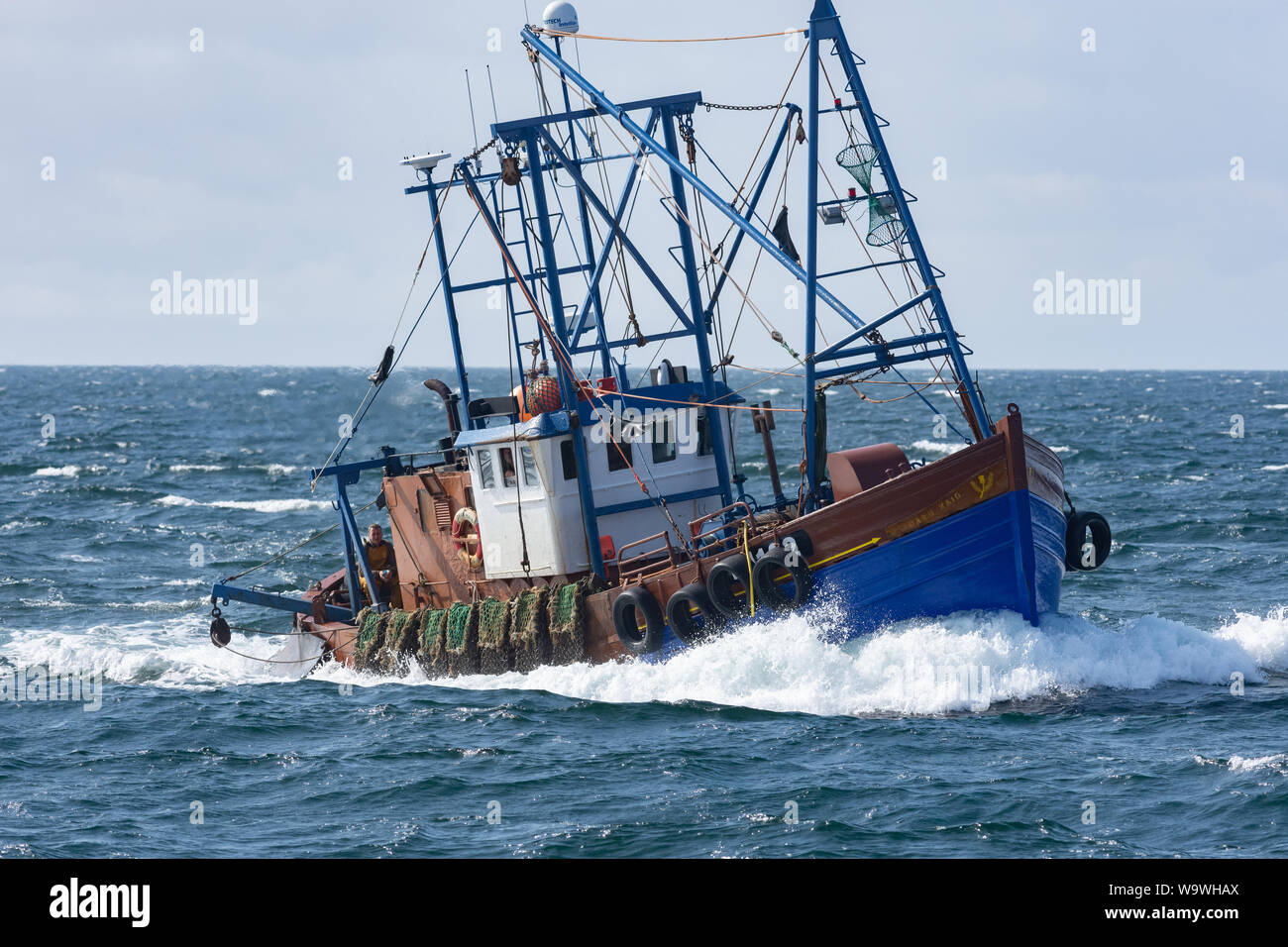 Scallop dredger boat ploughs through waves at sea with sunny skies behind, off the west coast of Scotland, with the dredges hanging off the side Stock Photo