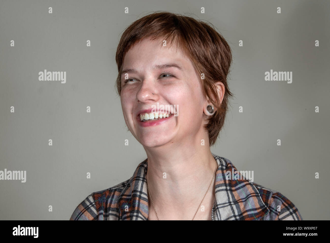 A pretty young woman in a casual flannel shirt looks amused,  in a head-and-shoulders studio shot. Stock Photo