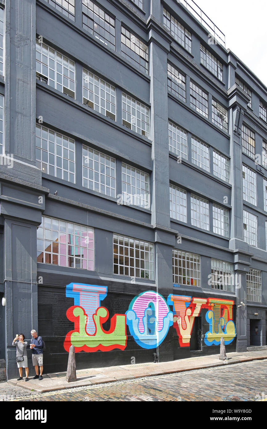 A couple stand by the word LOVE painted onto an industrial building in Ebor Street in Shoreditch, London, UK. Brightly coloured,1960's syle lettering. Stock Photo