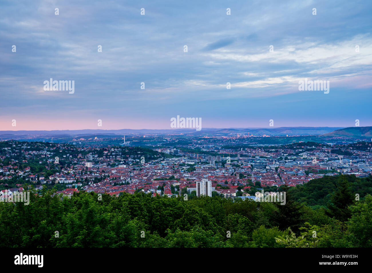 Germany, Above green treetops and red roofs of skyline of large city stuttgart from hilltop of mount rubble at sunset in summer Stock Photo