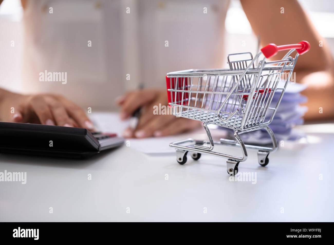 Close-up Of Person Calculating Bill With Calculator Near Shopping Cart On Desk Stock Photo