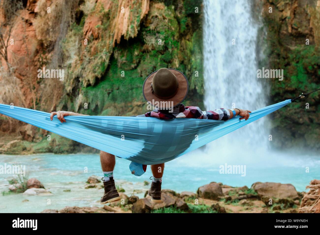 Wide shot of a male lying on a hammock beside a waterfall flowing down from a hill Stock Photo