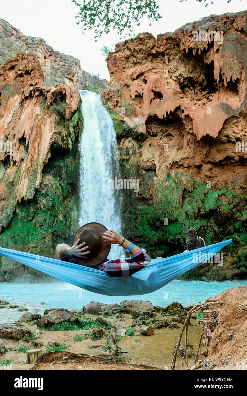 Vertical shot of a male lying on a hammock beside a waterfall flowing down from a hill Stock Photo