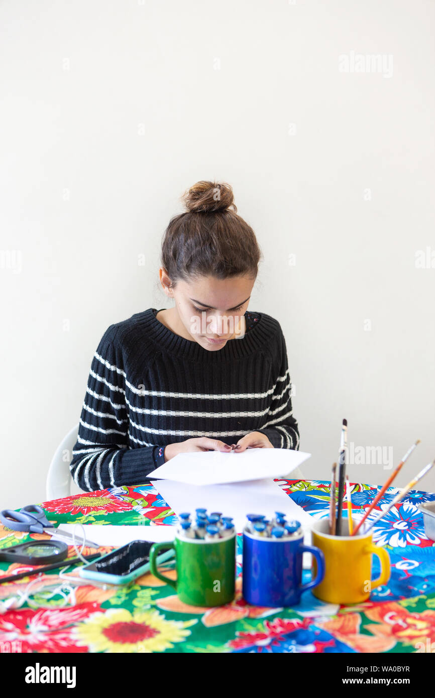 Young talented woman artist sitting at the table with flowered tablecloth making art with pencils, paper, scissors, magnifying glass, brush and paints Stock Photo