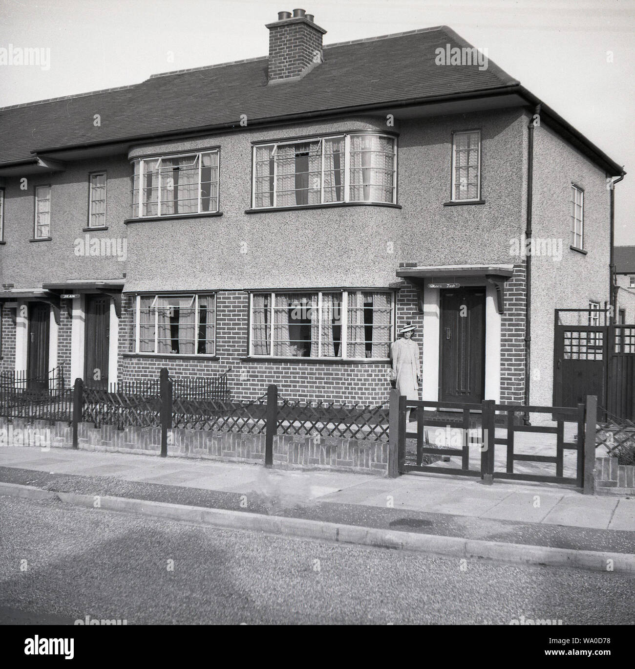 1950s, historical, lady standing outside the entrance of a 1930s built semi-detached house, with a pebbledash or roughcast covering on the upper leve and side wall, London, England, UK. This plaster coating on the outside walls was often applied to houses built in the 1920s and 30s for reasons of design and style and added rudimentary weather protection, but decllined in popularity in later eras. Stock Photo