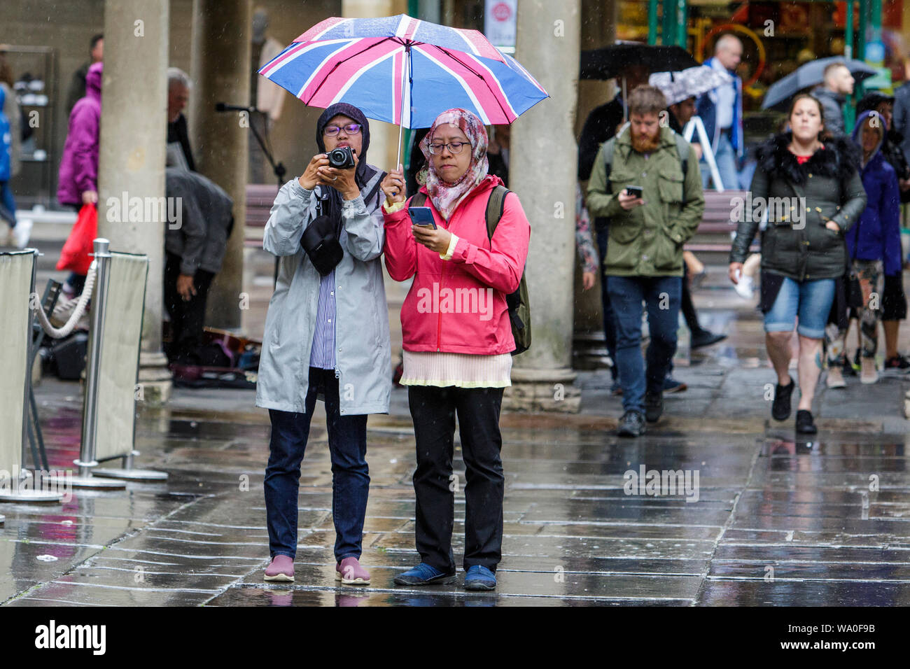 Bath, Somerset, UK. 16th August, 2019. Tourists are pictured as they take a photograph of Bath Abbey as heavy rain showers make their way across the UK.  Credit: Lynchpics/Alamy Live News Stock Photo