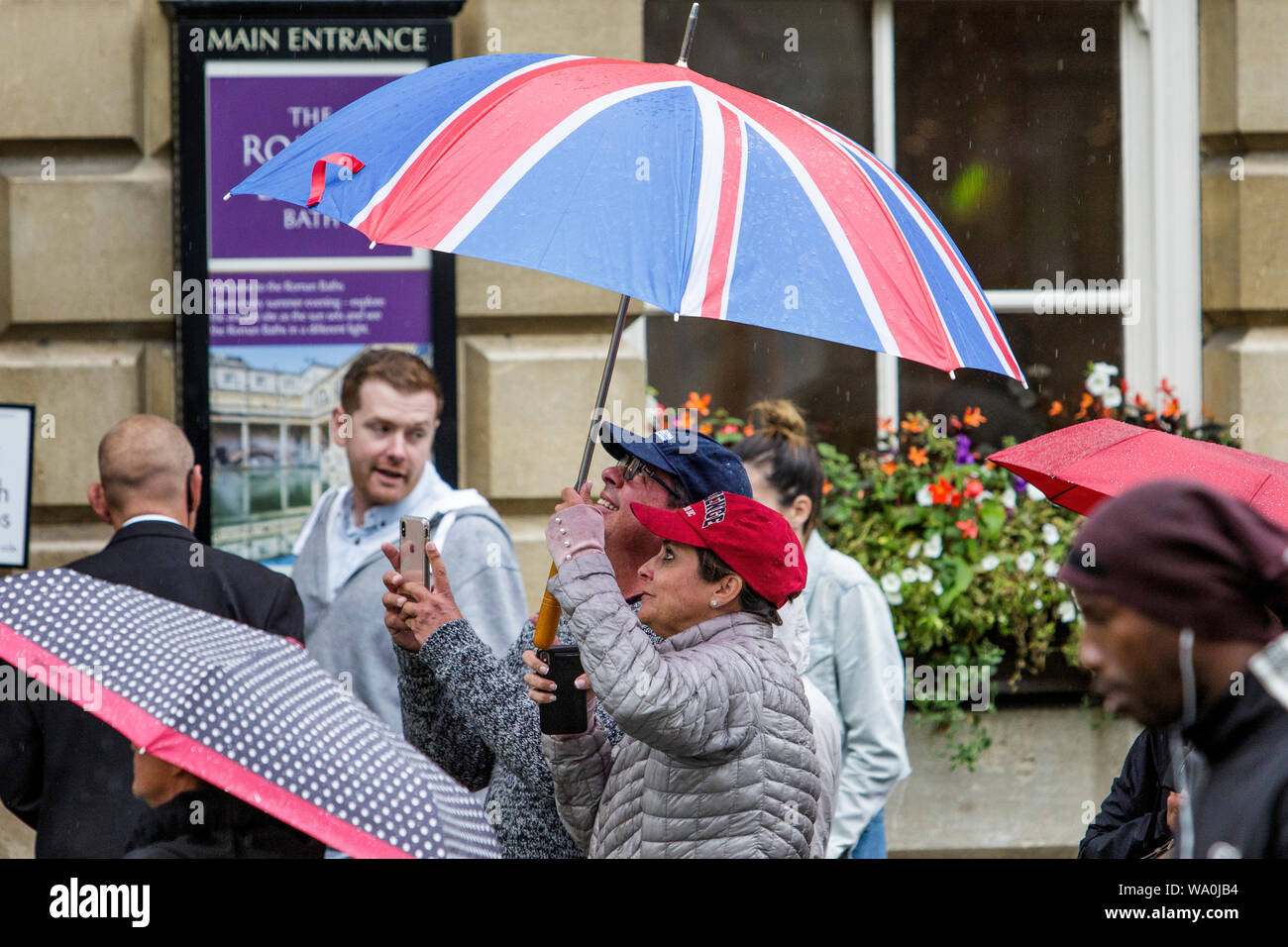 Bath, Somerset, UK. 16th August, 2019. Tourists are pictured as they take a photograph of Bath Abbey as heavy rain showers make their way across the UK.  Credit: Lynchpics/Alamy Live News Stock Photo