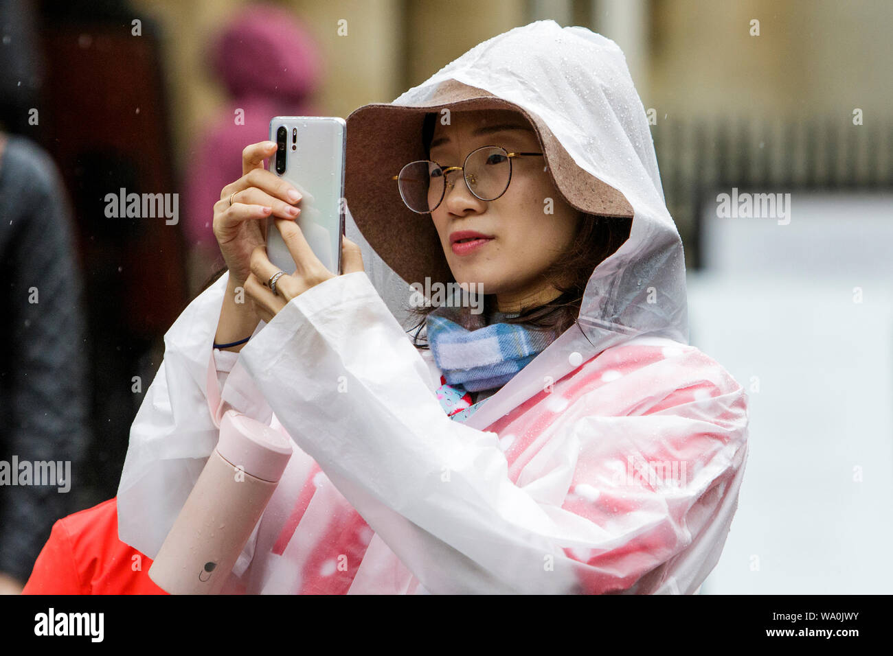 Bath, Somerset, UK. 16th August, 2019. A tourist is pictured as she takes a photograph of Bath Abbey as heavy rain showers make their way across the UK.  Credit: Lynchpics/Alamy Live News Stock Photo