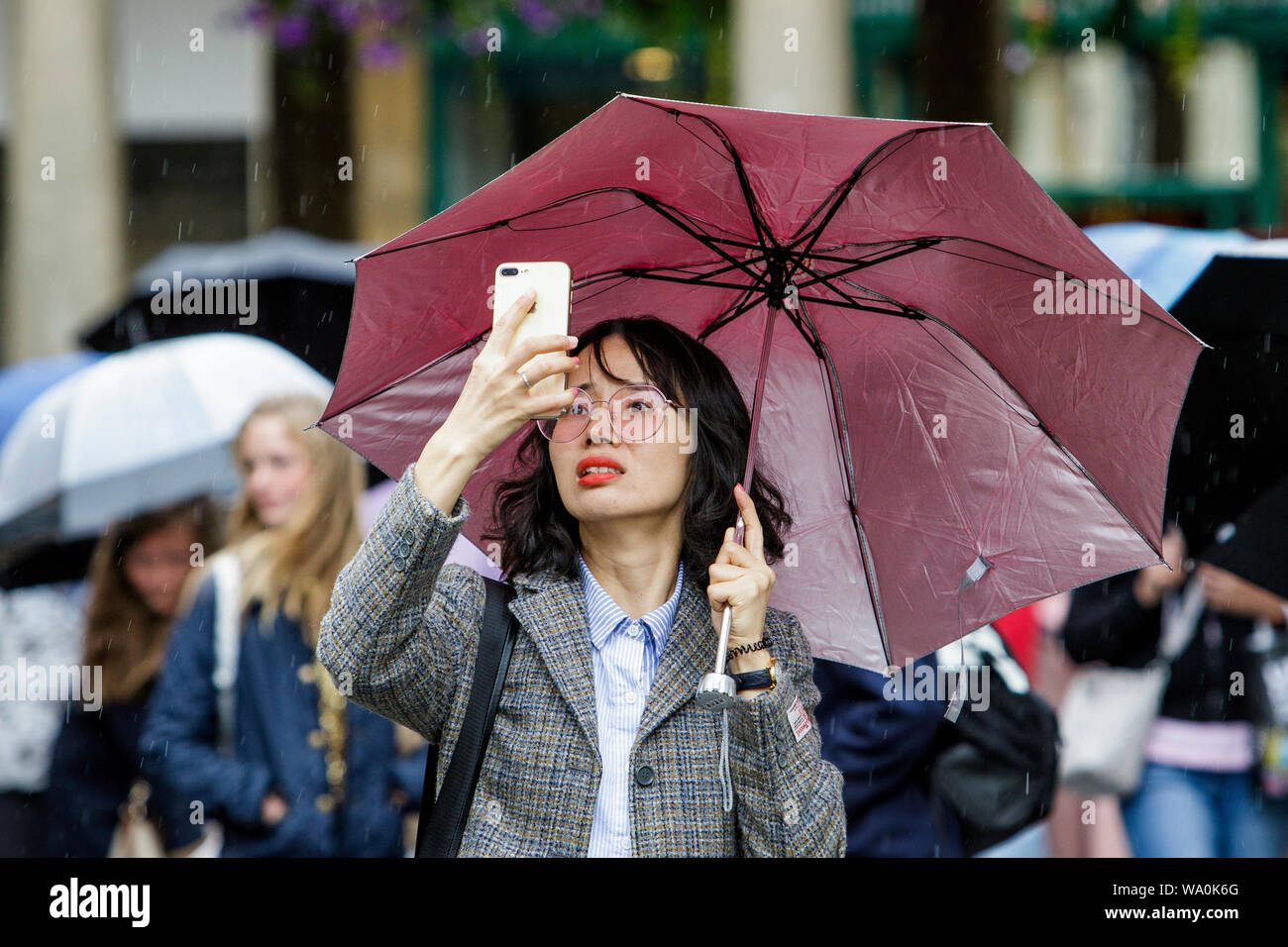 Bath, Somerset, UK. 16th August, 2019. A tourist is pictured as she takes a photograph of Bath Abbey as heavy rain showers make their way across the UK.  Credit: Lynchpics/Alamy Live News Stock Photo