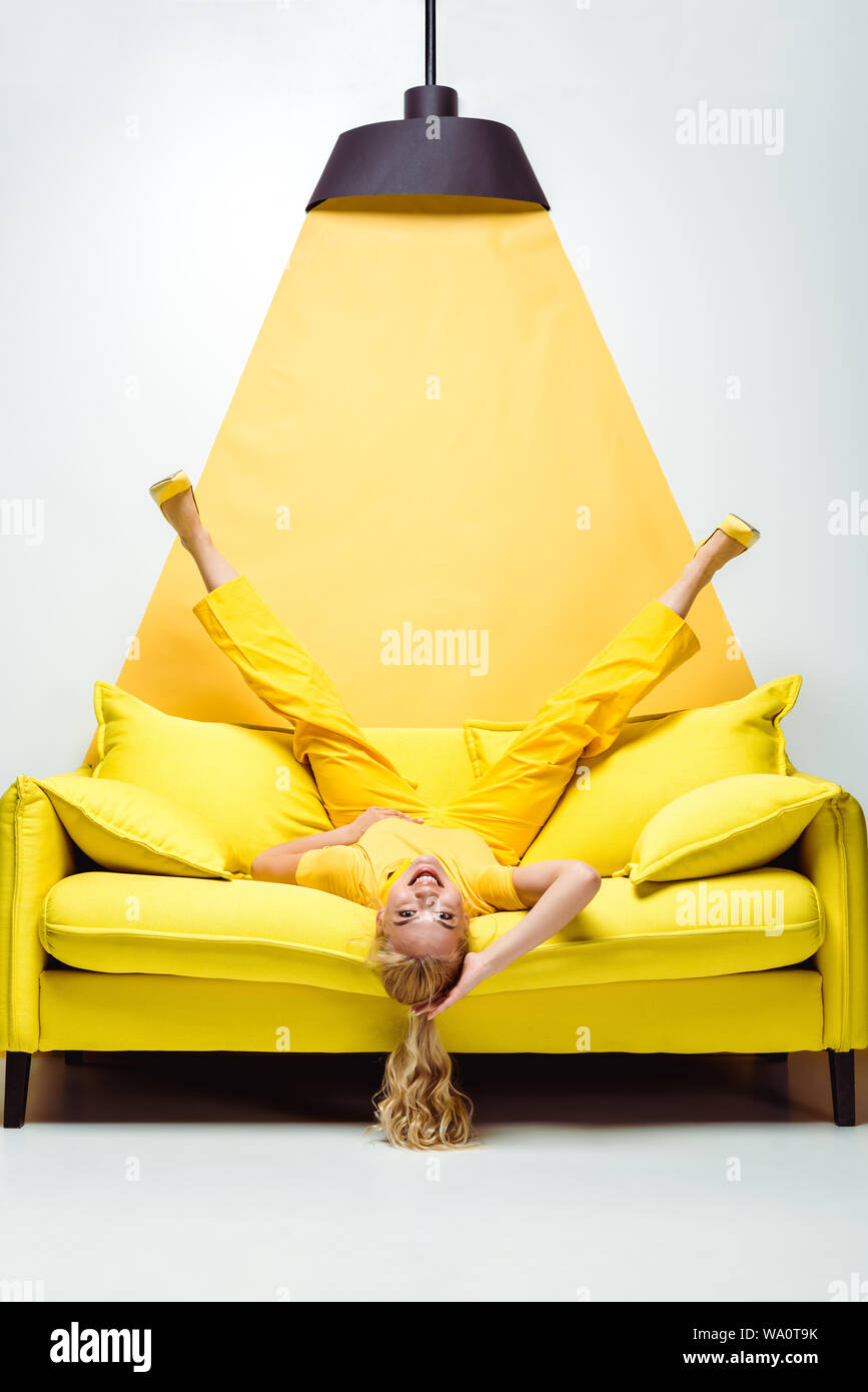 happy blonde woman lying upside down on sofa on white and yellow Stock