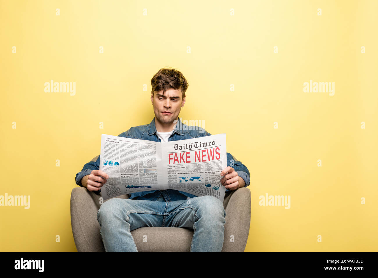 serious young man sitting in armchair and reading newspaper with fake ...