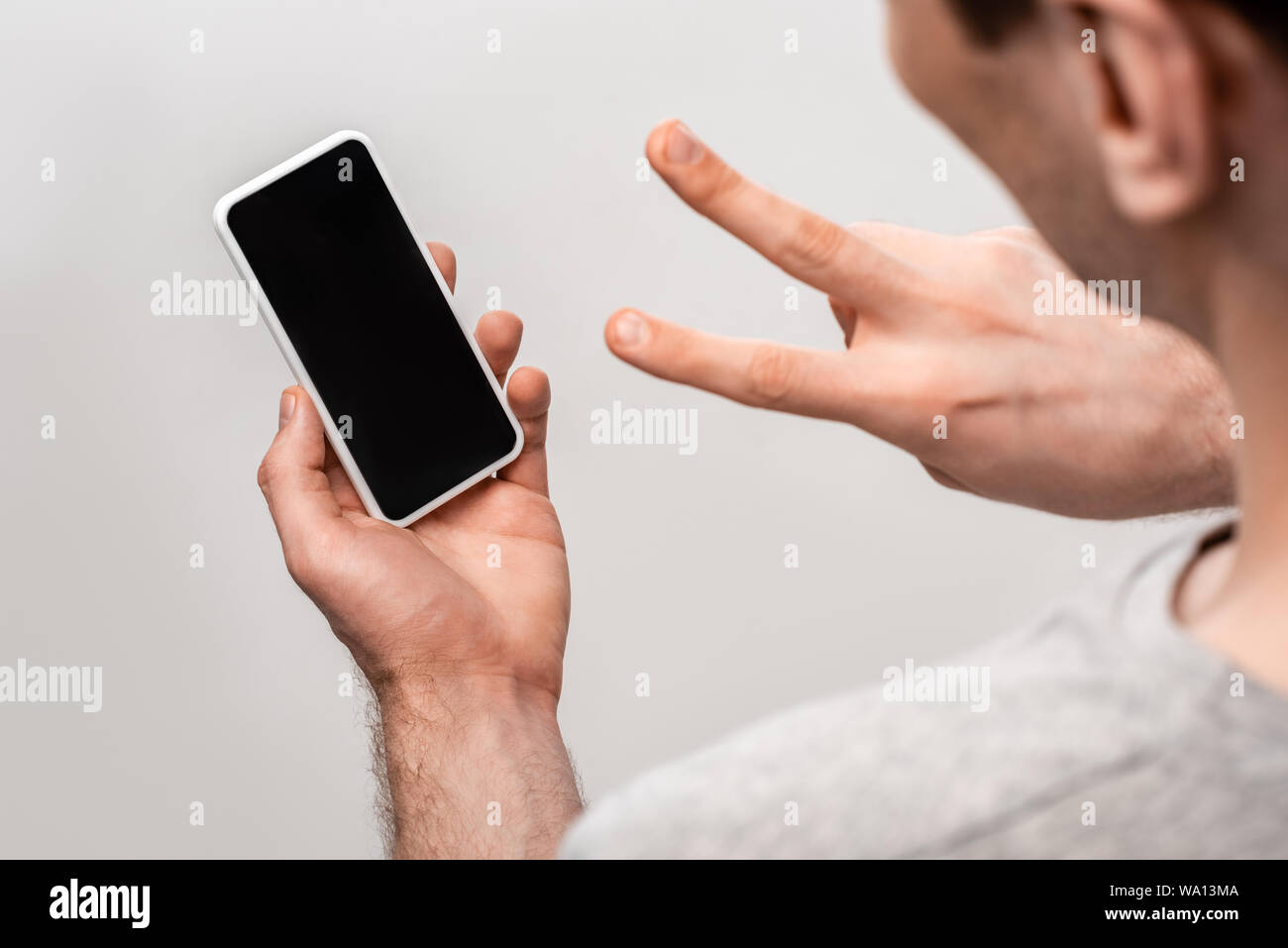 cropped veiw of man showing victory sign while holding smartphone with blank screen isolated on grey Stock Photo