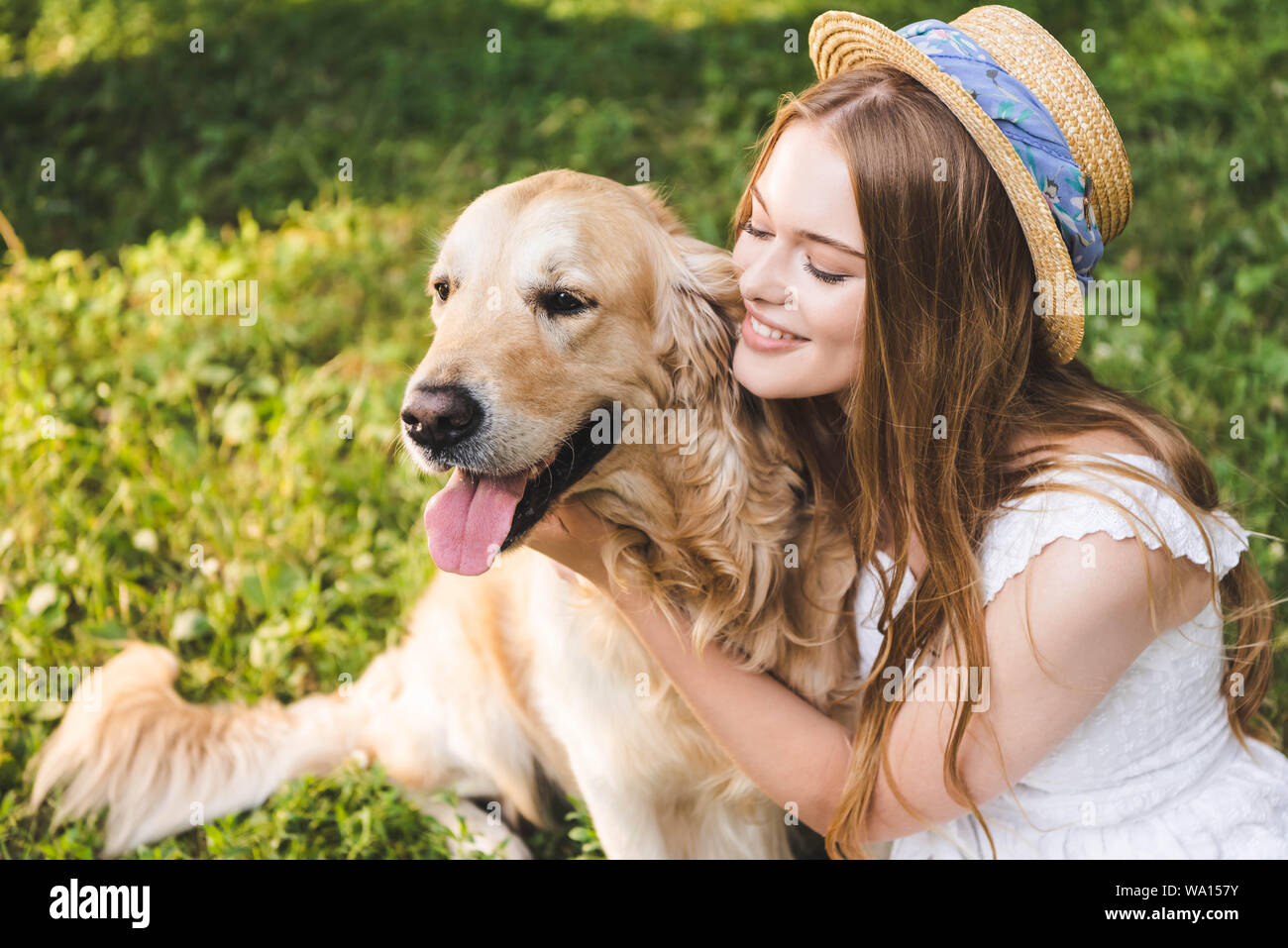 beautiful girl in white dress and straw hat hugging golden retriever while  sitting on meadow and looking at dog Stock Photo - Alamy
