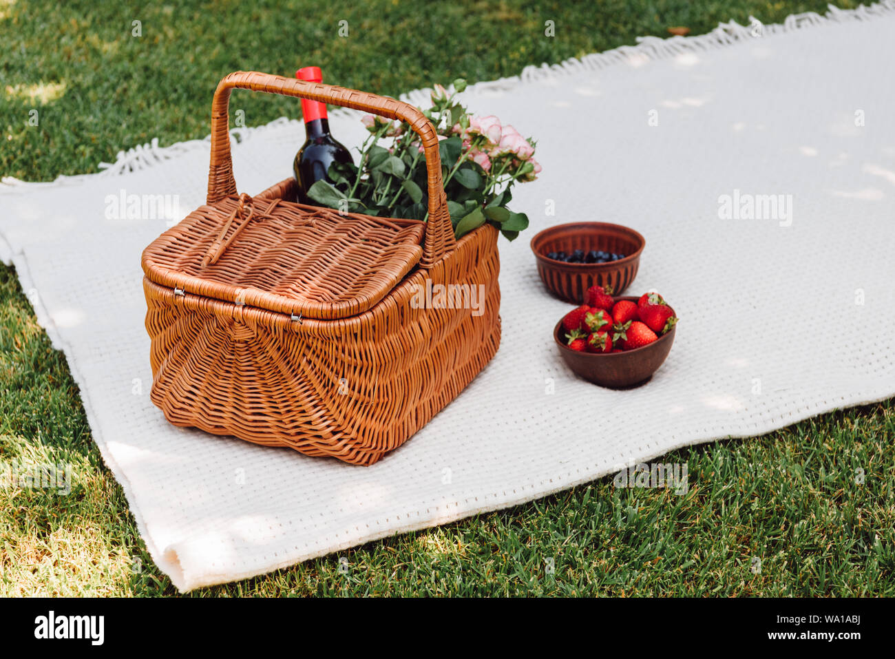 wicker basket with roses and bottle of wine on white blanket near strawberries in shadow in garden Stock Photo
