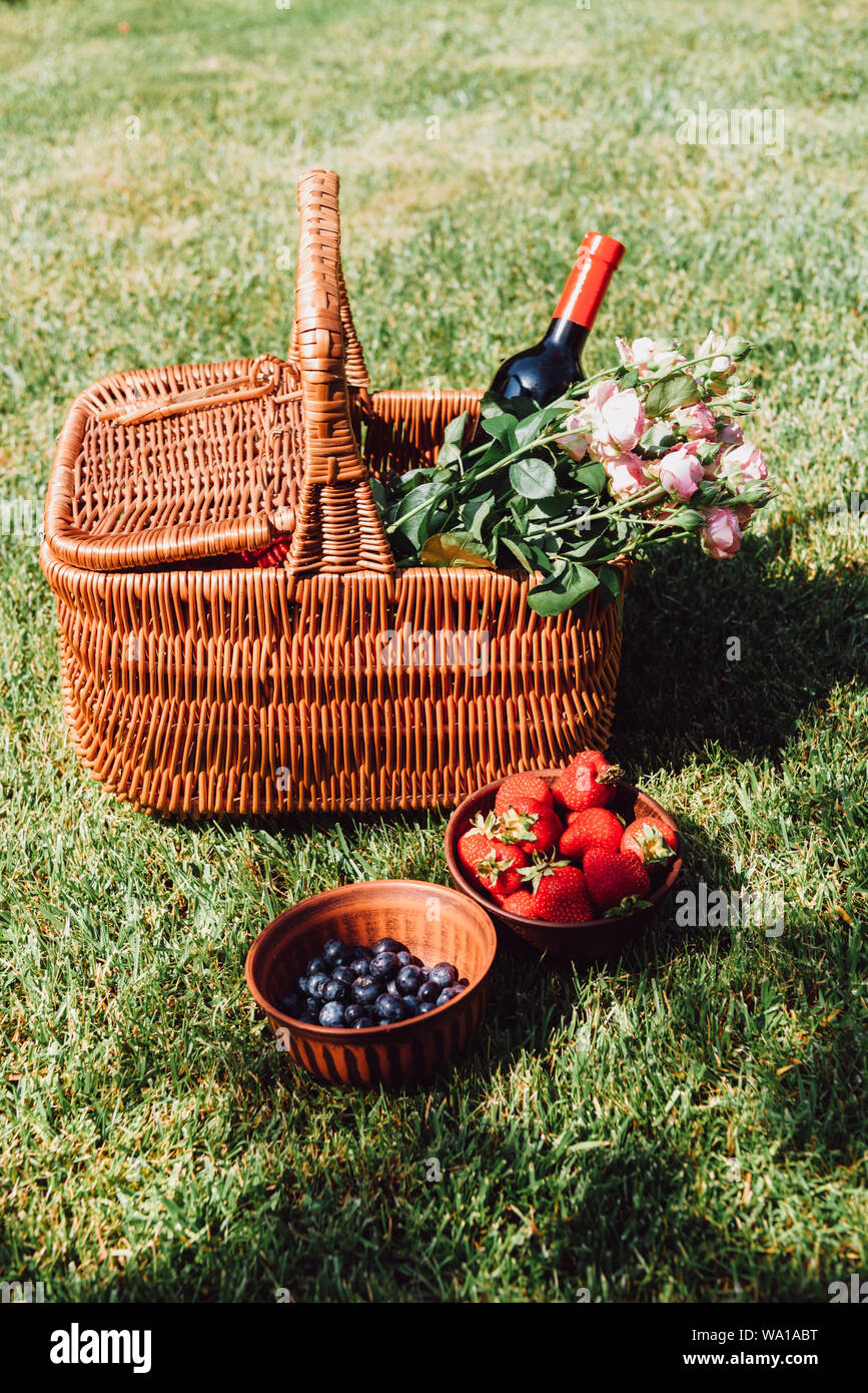 wicker basket with roses and bottle of wine on green grass near strawberries and blueberries in bowls Stock Photo