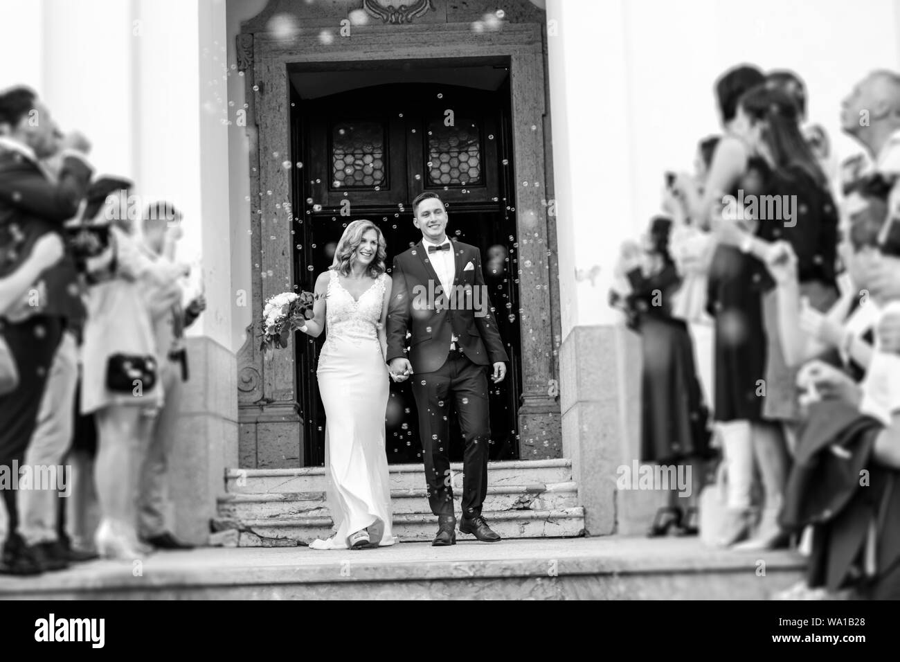 Newlyweds exiting the church after the wedding ceremony, family and friends celebrating their love with the shower of soap bubbles, custom undermining Stock Photo