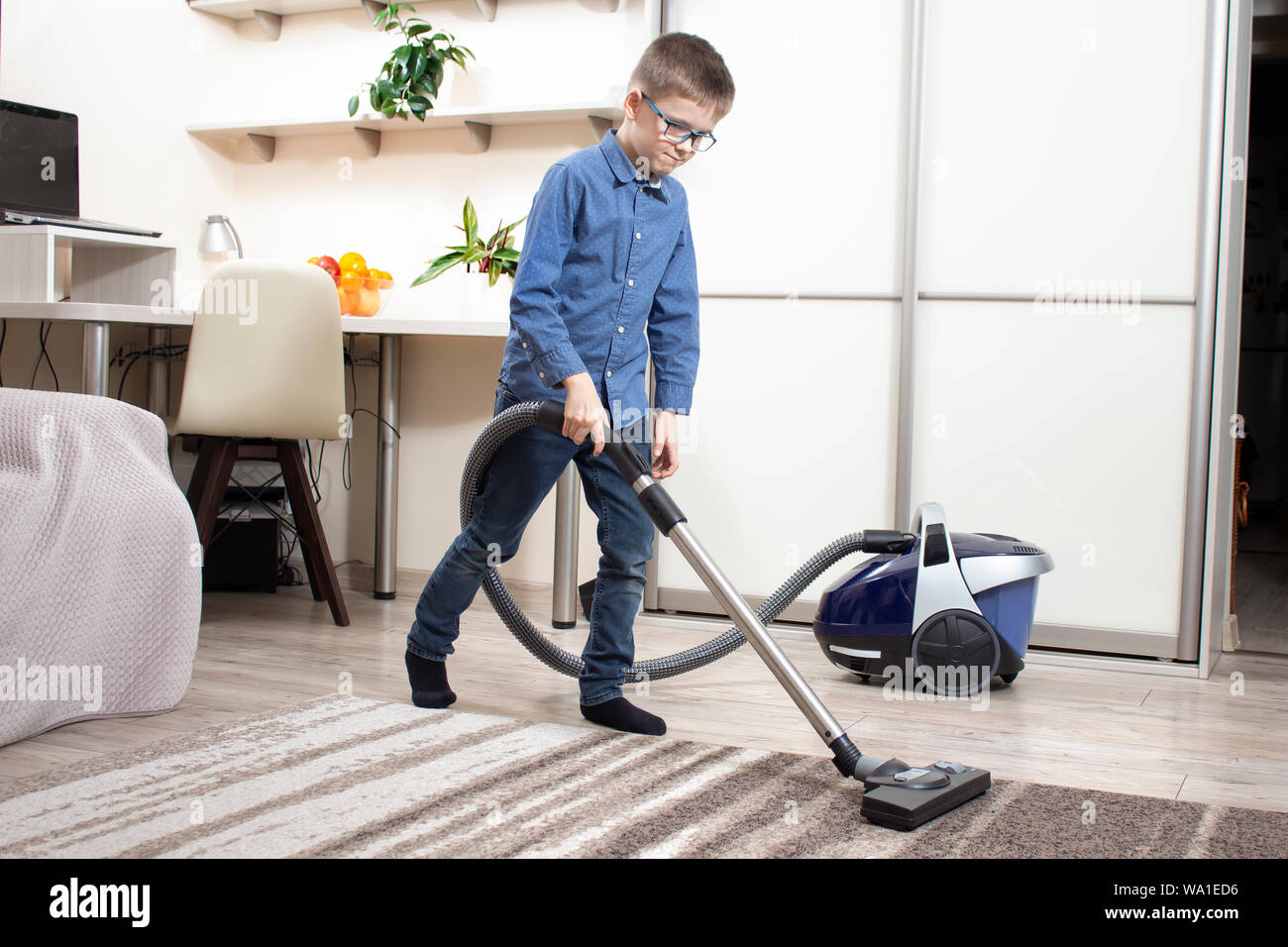 Vacuuming a rug in an apartment by a boy dressed in a blue shirt and jeans pants. Stock Photo