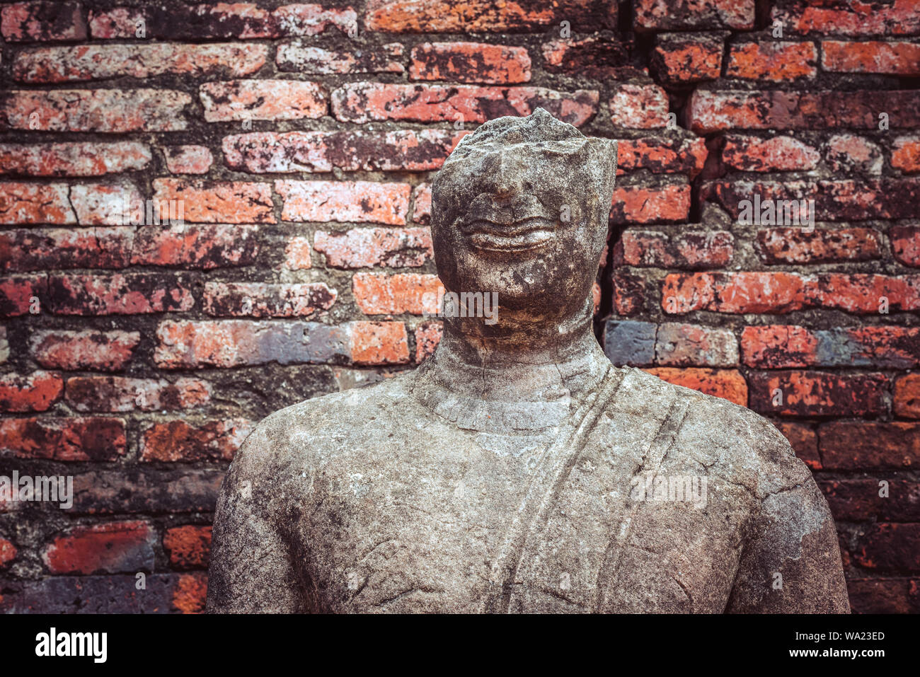 Buddhist smile statue. Crumbling statue with broken-off upper half of the head (without eyes) on the shabby brick wall background. Ayutthaya, Thailand Stock Photo