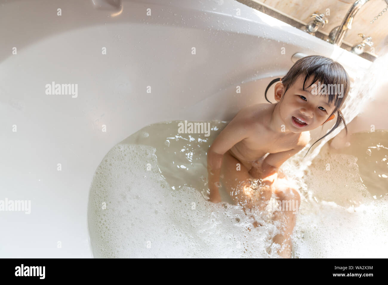 Asian happy laughing kid girl taking a bath playing with foam bubbles. Little child in a bathtub. Smiling kid in bathroom with sunlight window backgro Stock Photo