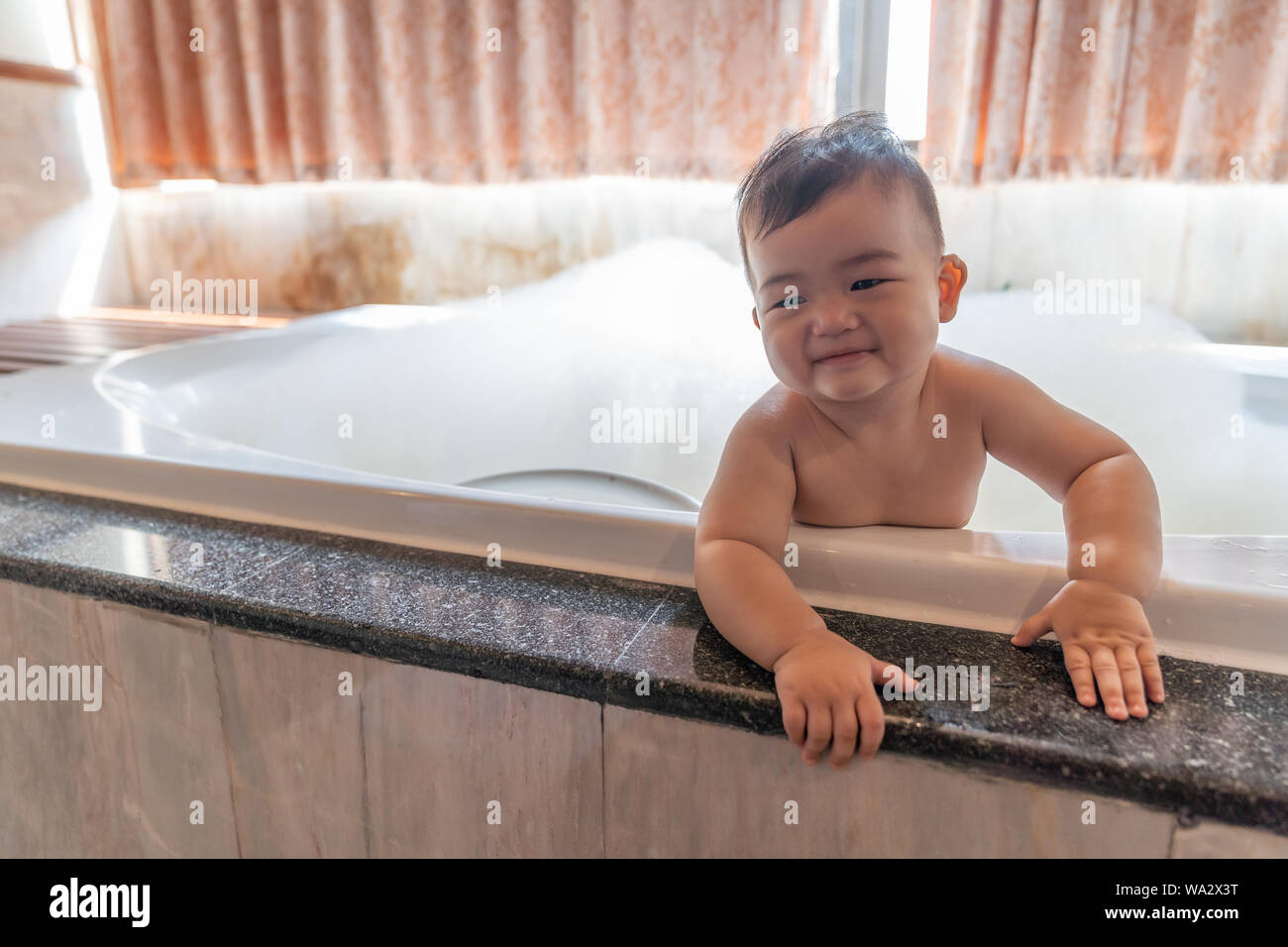 Asian happy laughing baby boy taking a bath playing with foam bubbles. Little child in a bathtub. Smiling kid in bathroom with sunlight window backgro Stock Photo