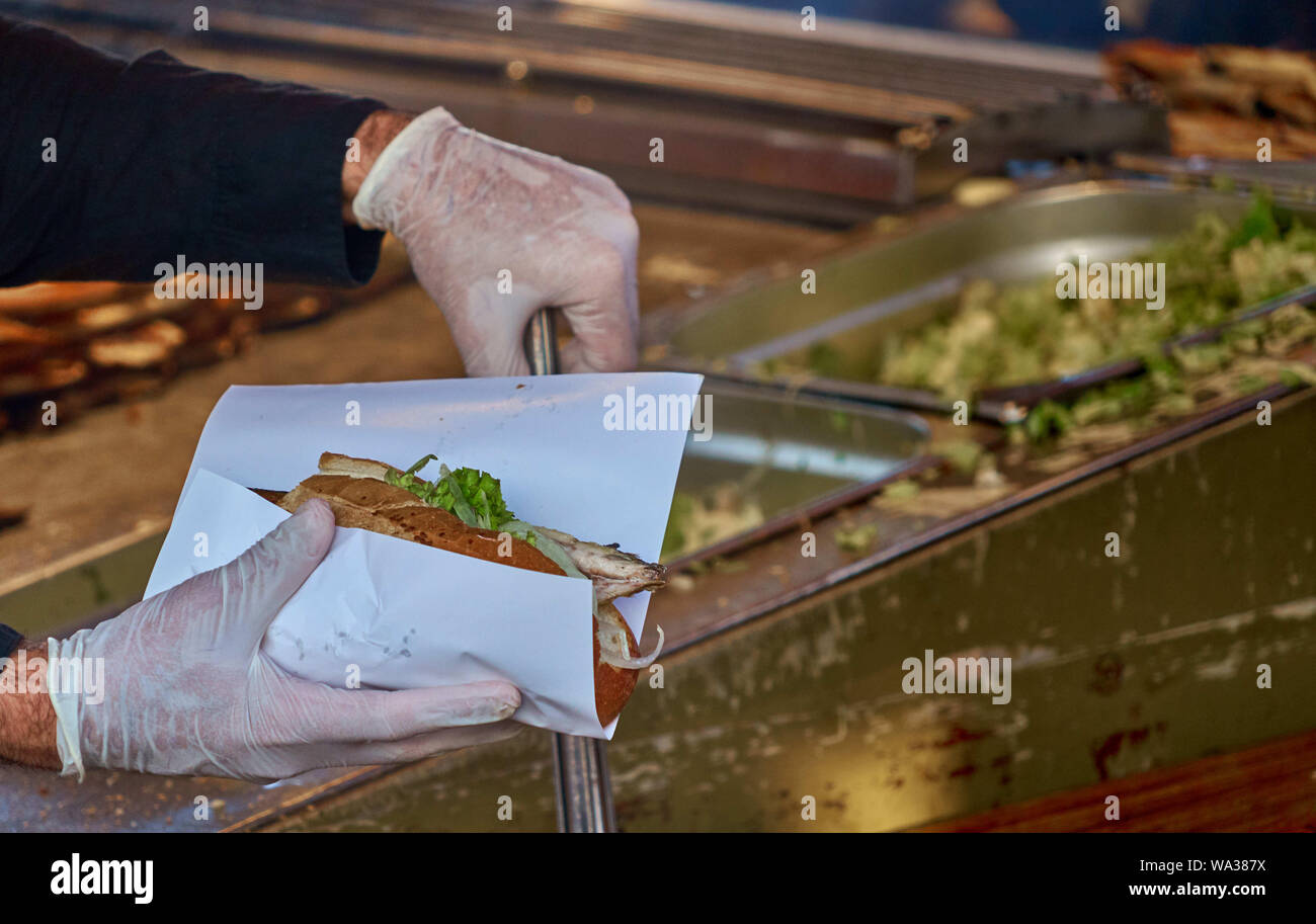 Chef with hygienic rubber gloves and a pair of tongs prepares a fish roll from a roll, a piece of mackerel fillet and a salad with onions. Stock Photo
