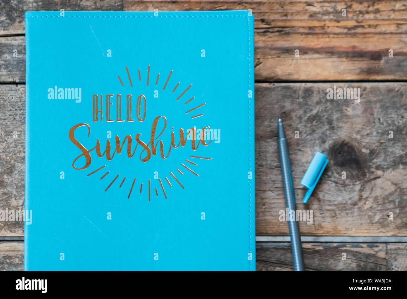 Overhead shot of a 'hello sunshine' blue textbook with a ball-point pen next to it on wooden surface Stock Photo