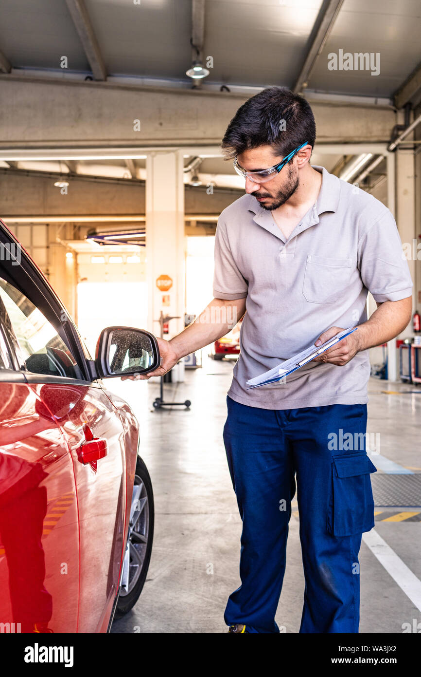 Technician with safety glasses inspecting with the hand the right rearview of a red car in a