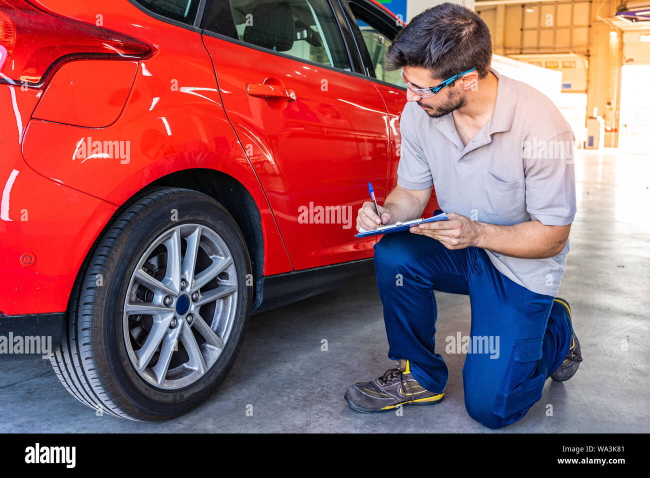 Technician with safety glasses checking the tires of a red car during a vehicle inspection Stock