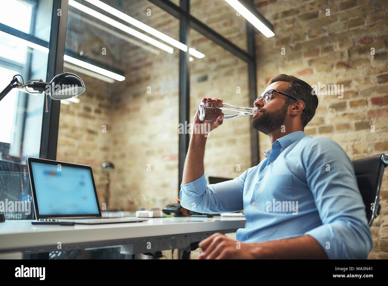 So thirsty. Side view of young bearded businessman in eyeglasses and formal wear drinking fresh water while working in the modern office. Business concept. Work concept Stock Photo