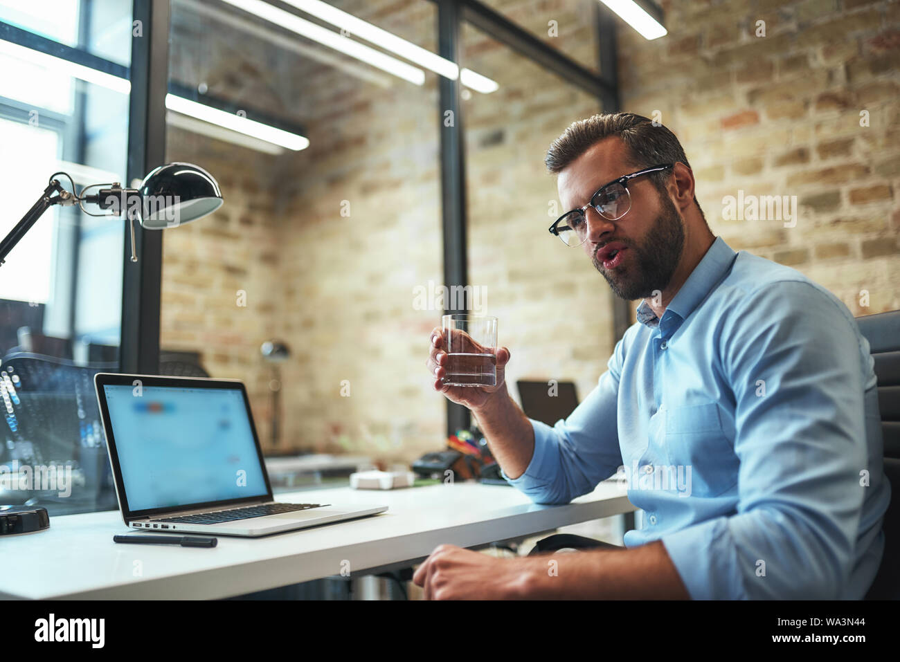 Refreshing. Thirsty bearded businessman in eyeglasses and formal wear drinking fresh water while working in the modern office. Business concept. Work concept Stock Photo