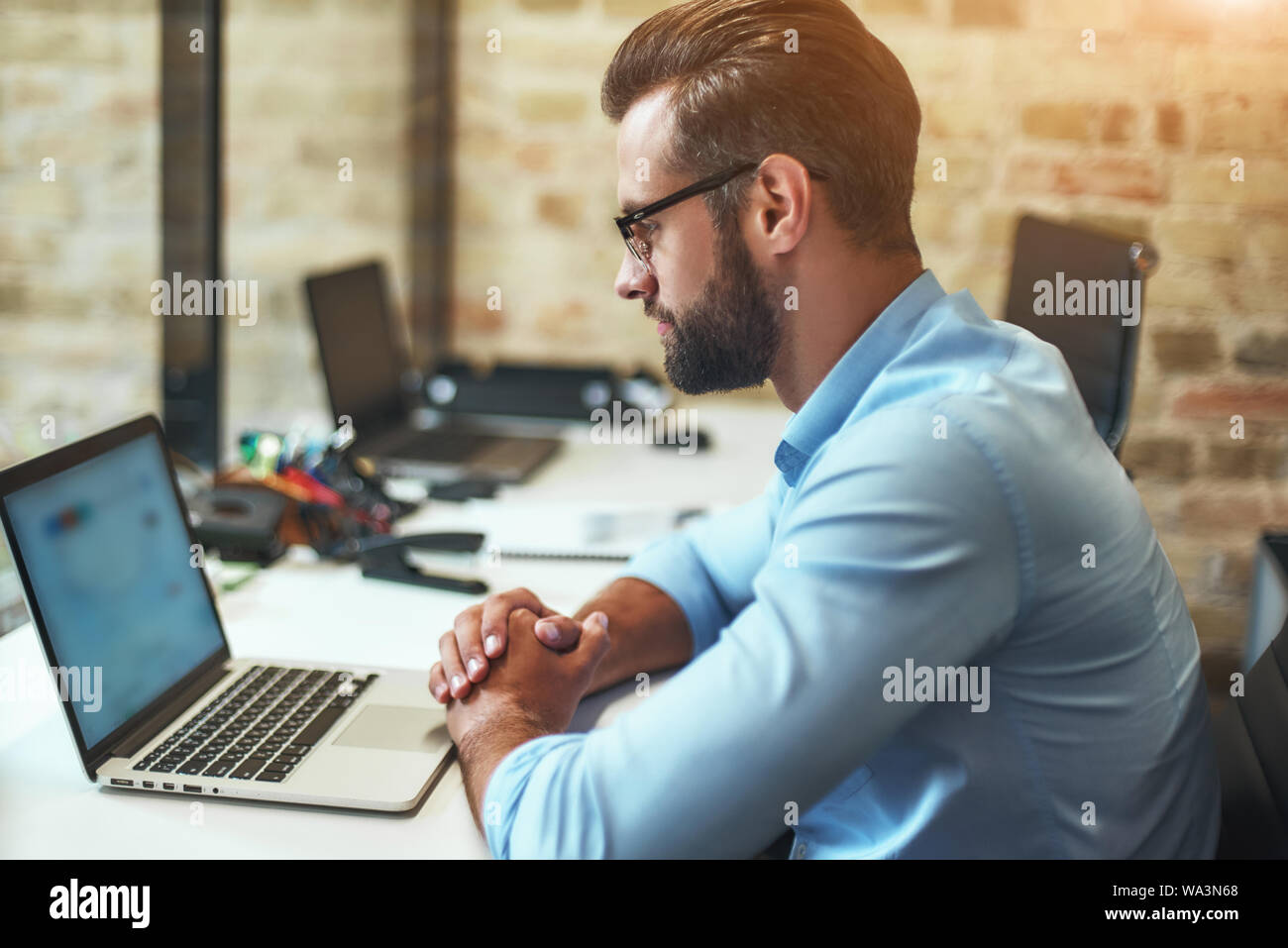 Analyzing reports. Side view of young bearded businessman in eyeglasses and formal wear looking at laptop while sitting in the modern office. Business concept. Work concept Stock Photo