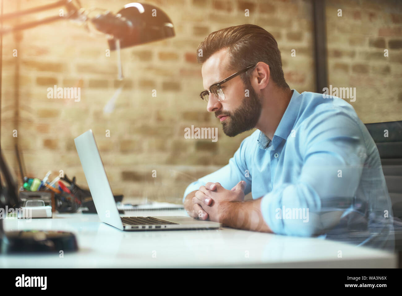 Analyzing reports. Side view of young bearded businessman in eyeglasses and formal wear looking at laptop while sitting in comfortable office. Business concept. Work concept Stock Photo