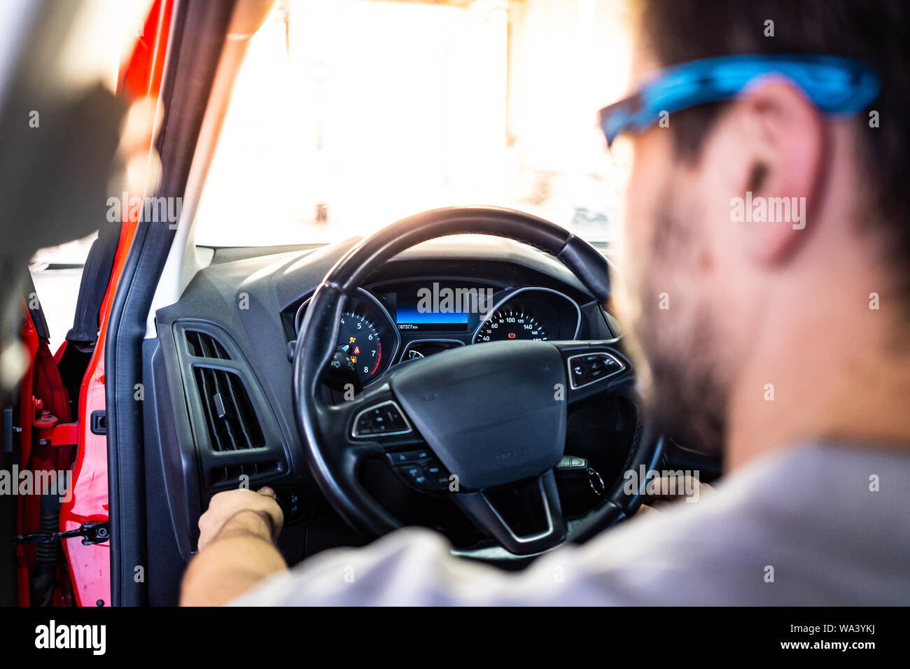 Technician with safety glasses starting the engine of a red car during a vehicle inspection