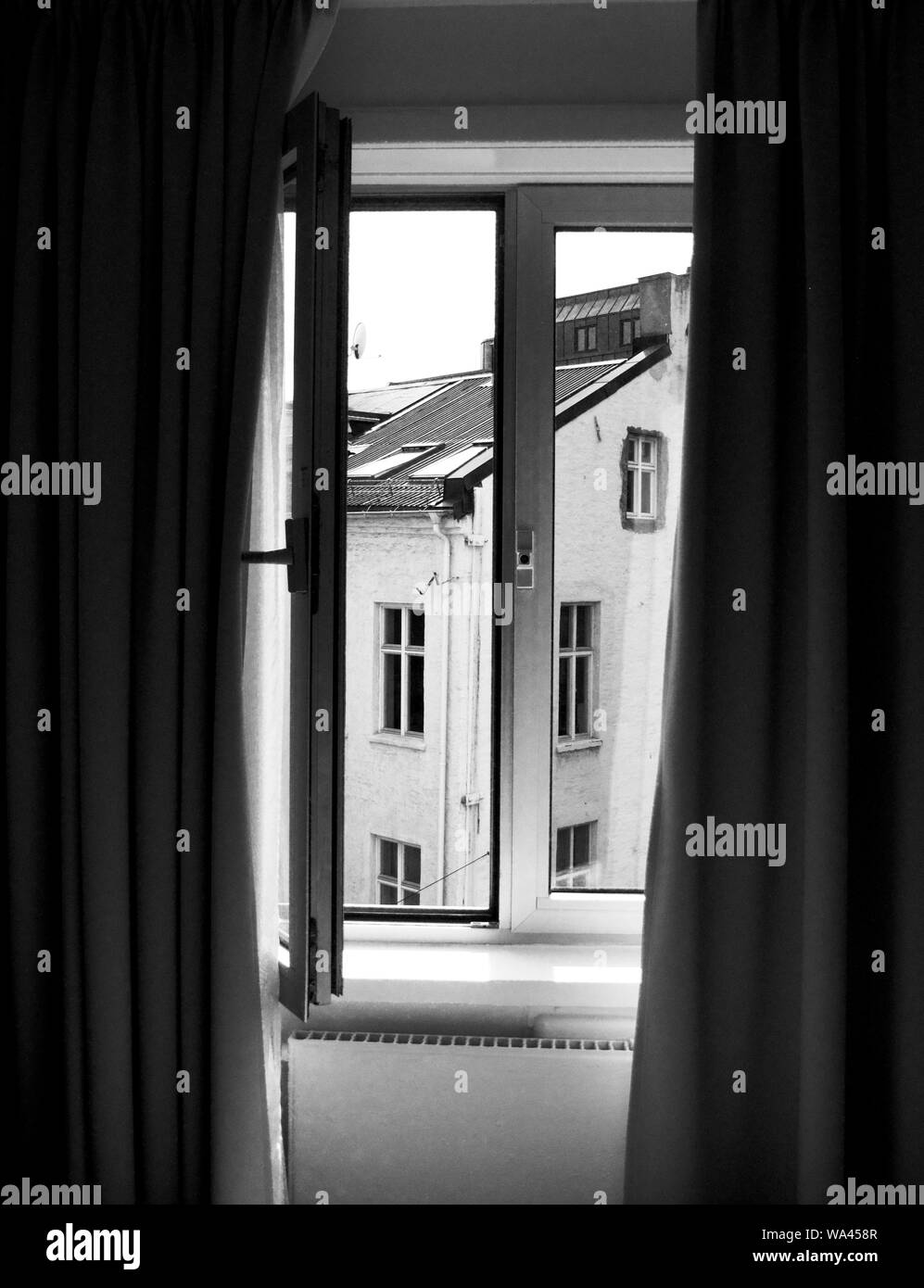 Vertical shot of an open window near curtains with a view of a house in black and white Stock Photo