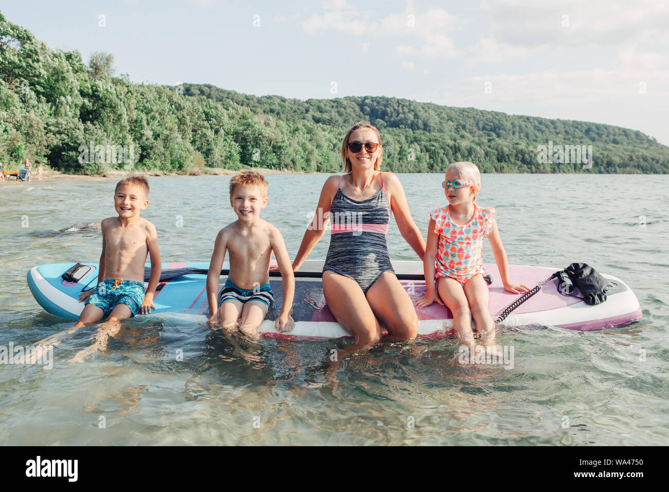 Caucasian woman parent sitting on paddle sup surfboard in water with kids children. Modern outdoor family activity. Summer aquatic activity Stock Photo