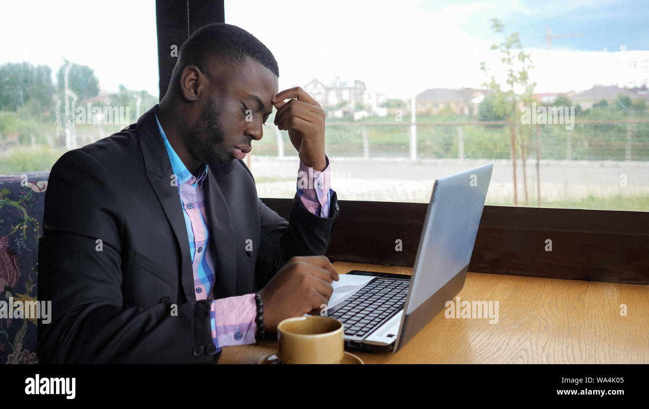 Afro american businessman tries to type on computer sitting in cafe. Black man works on laptop but it's broken and he is nervous and knocking on the keys. He has a computer problems. Battery is low. Stock Photo