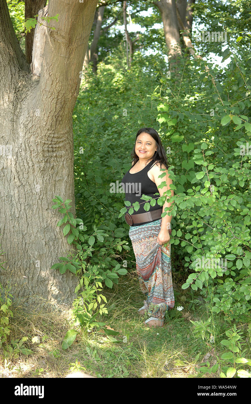 A lovely young woman in a long dress standing in the forest on a big tree in sunshine, smiling Stock Photo