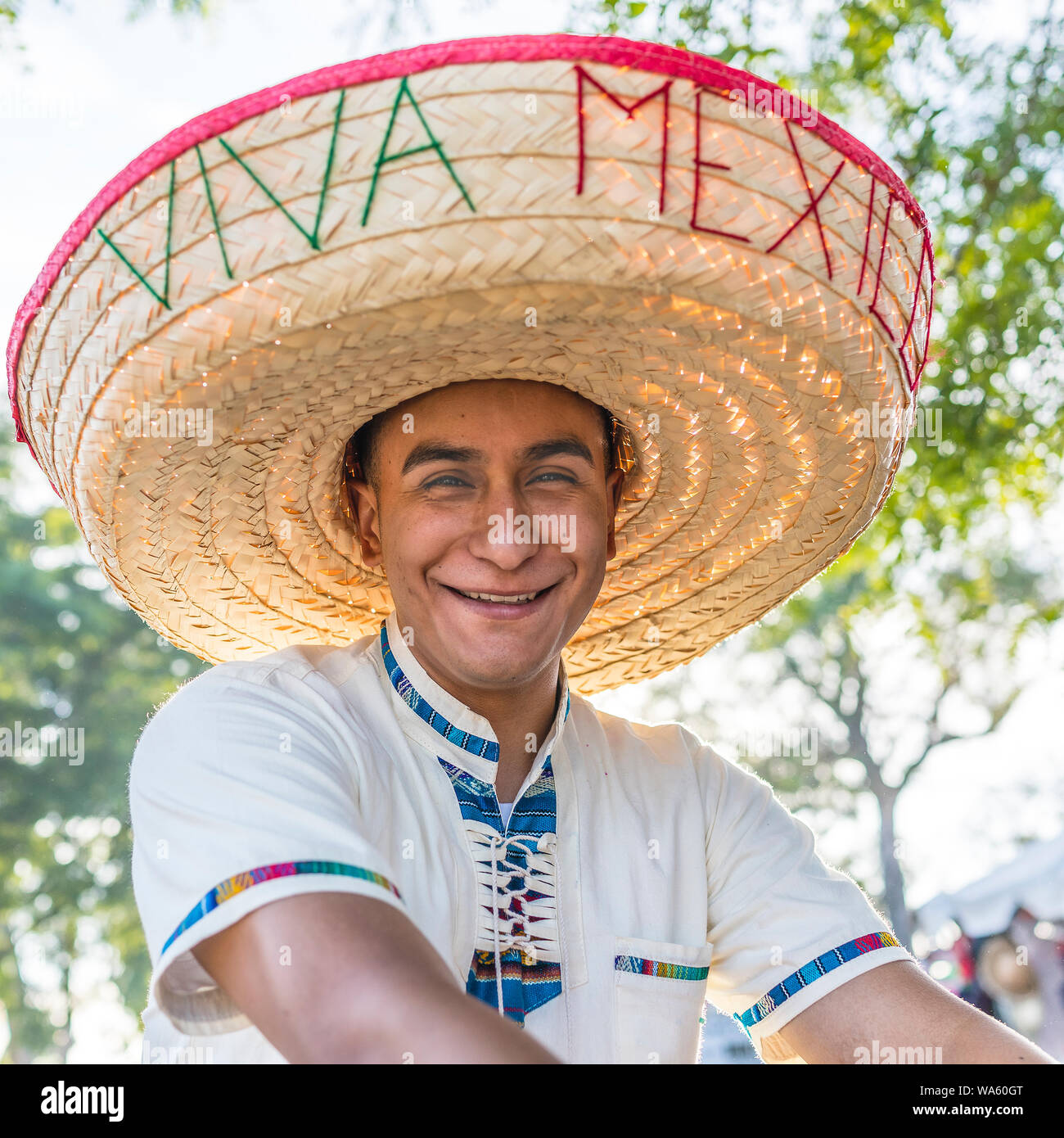 A 20 something HIspanic male wears and oversized straw sombrero hat with 'Viva Mexico' stitched on it in yarn and smiles while facing forward. Stock Photo