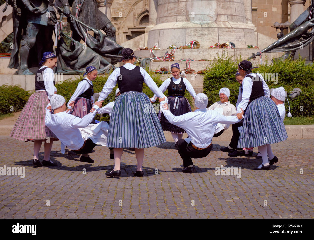 Hungarian Traditional dance troop in folkloric customs with a public performance in square. Dance in circle with man doing crunching dance. Stock Photo