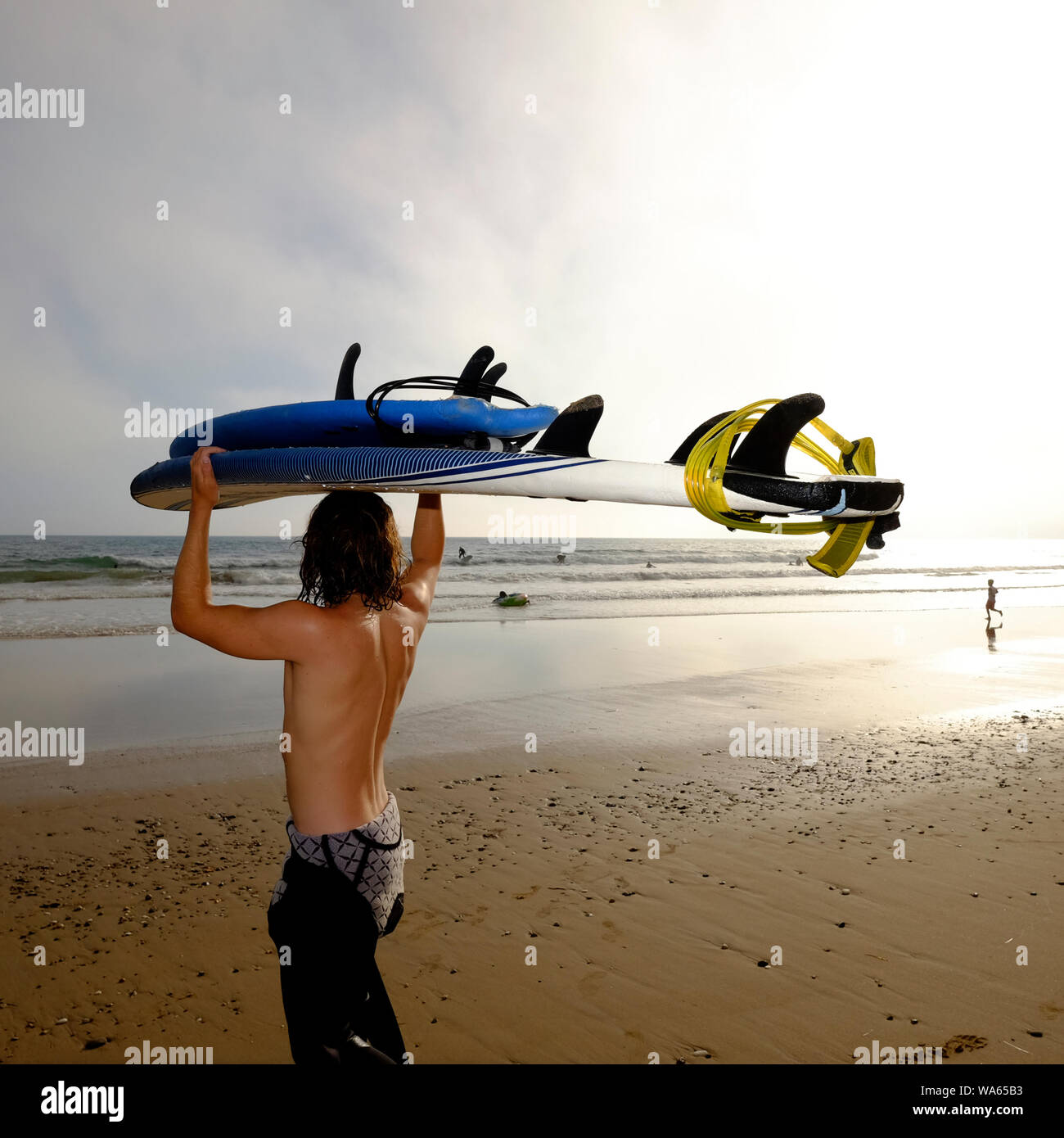 A young man carrying his surfboard on his head looking out into the sea on foot with the evening sun glowing brightly over Compton beach Stock Photo