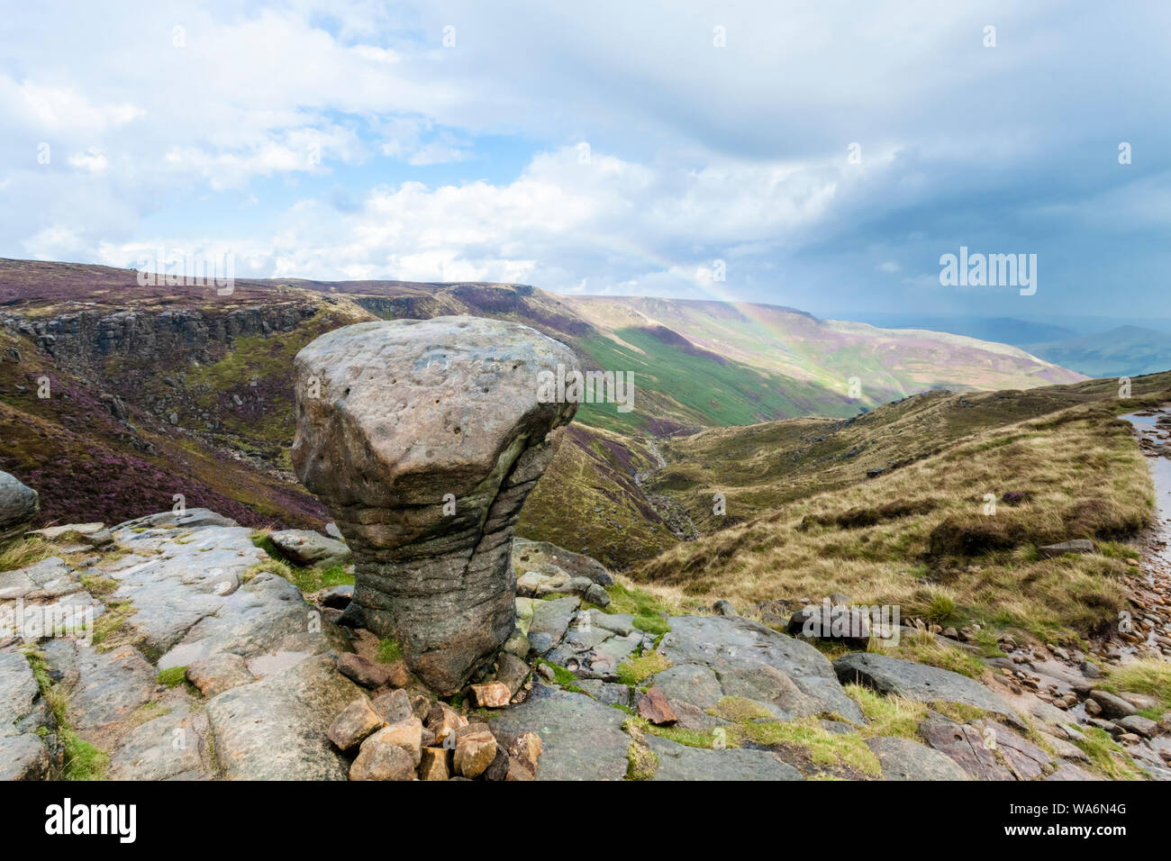 An eroded gritstone boulder at the top of Grindsbrook Clough, Kinder Scout after rain, with a rainbow in the distance. Derbyshire, England, UK Stock Photo