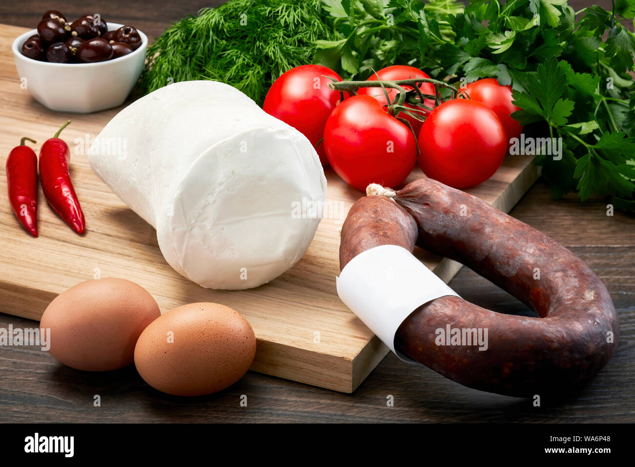 Turkish bryndza sheep milk cheese on wooden table with wooden cutting board with fermented sausage, eggs, olive, parsley and tomatoes. Stock Photo