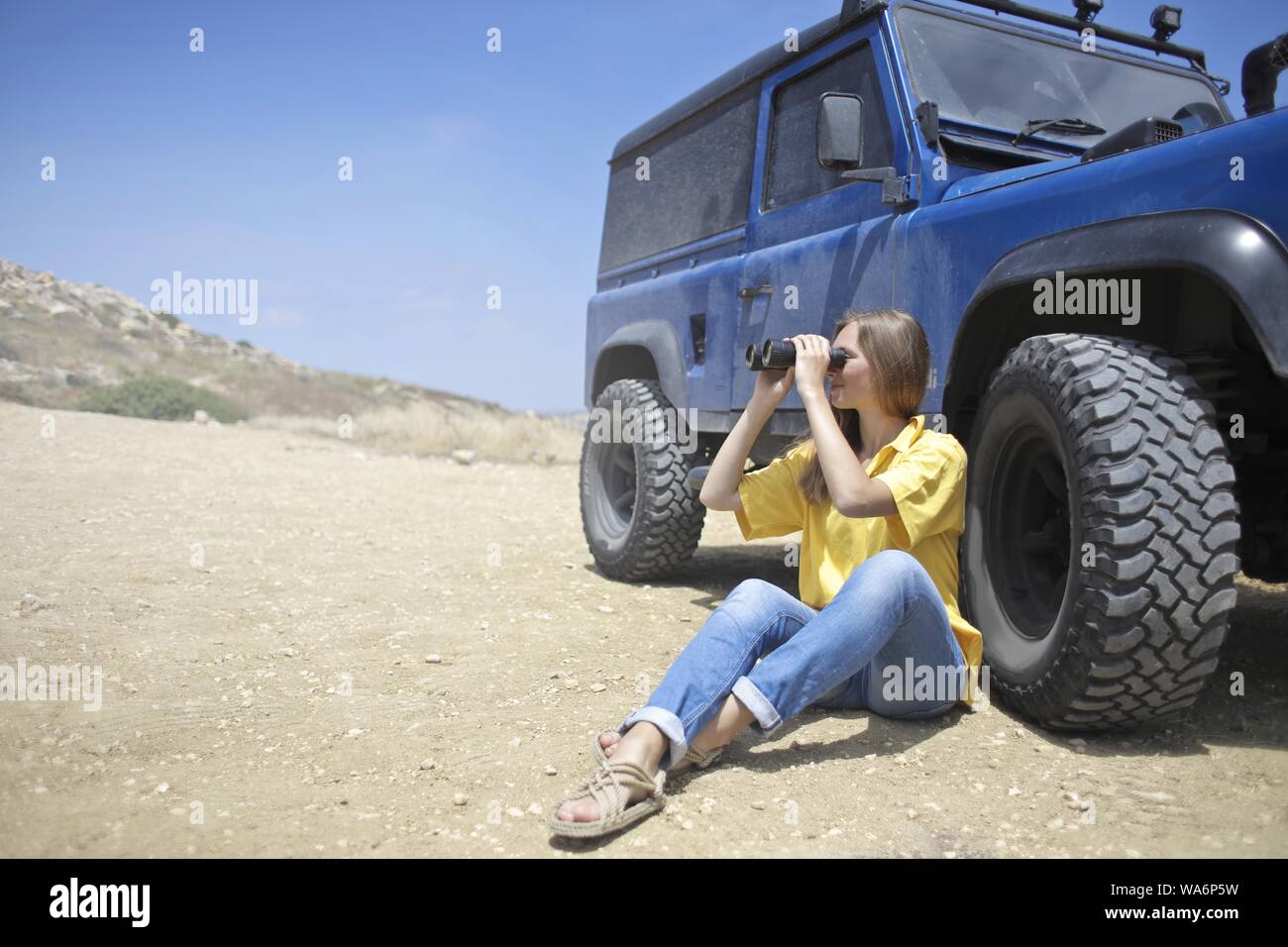 Shot Of A Woman Sitting on Soil Beside Blue Jeep Using Binoculars Stock Photo
