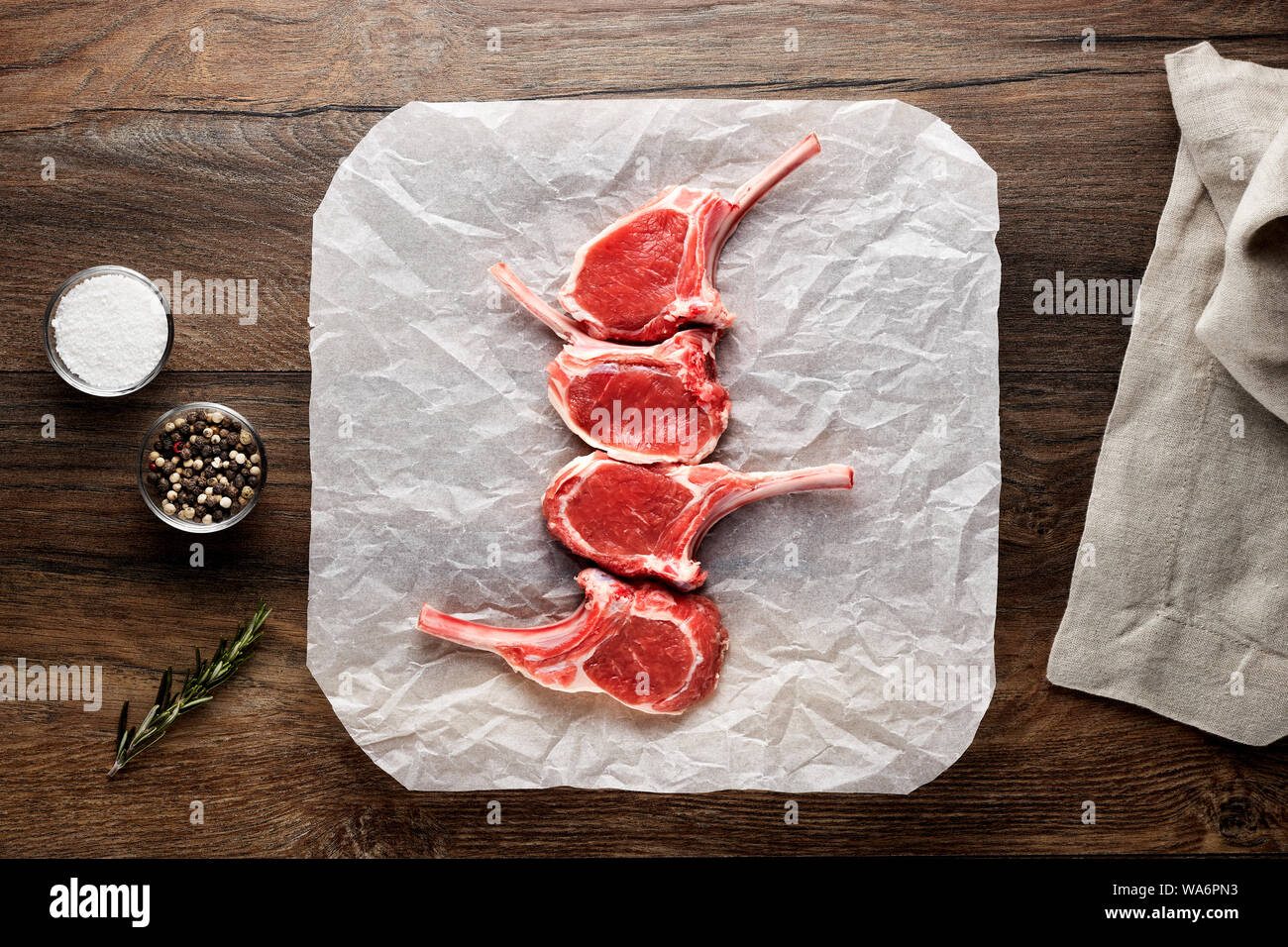 Slices of raw lamb rib chops on white cooking paper and wooden table. Decorated with salt, pepper, meat fork and napkin. Overhead view. Stock Photo