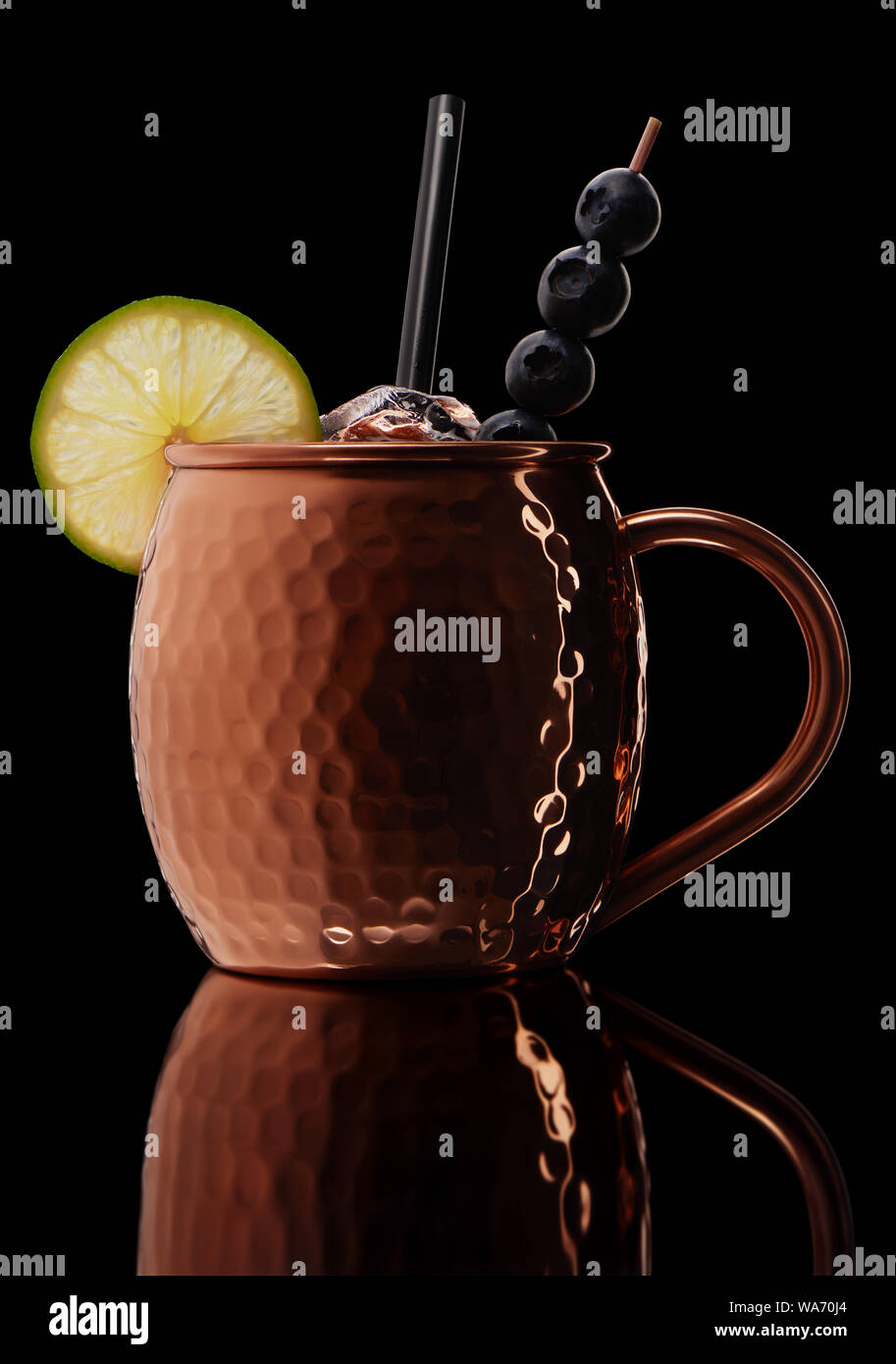 Icy cold Moscow mule cocktail with ginger beer, vodka, lime and blueberries in copper mug with reflection isolated on black background Stock Photo