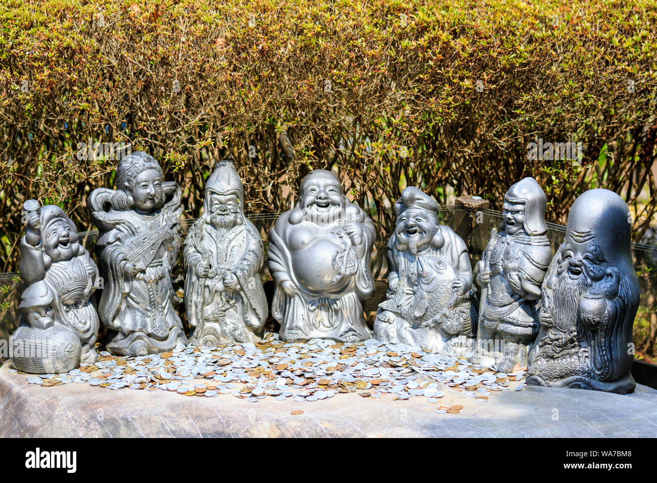 Japan, Miyajima. Daisho-in temple. Statues of the Japanese seven deities of good fortune outdoors in a line with pile of coins left as offerings. Stock Photo