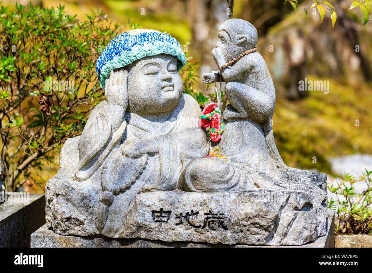 Japan, Miyajima. Daisho-in temple. Small Jizo statue of a reclining Buddhist monk wearing a knitted wool hat, and a monkey sitting next to him eating. Stock Photo