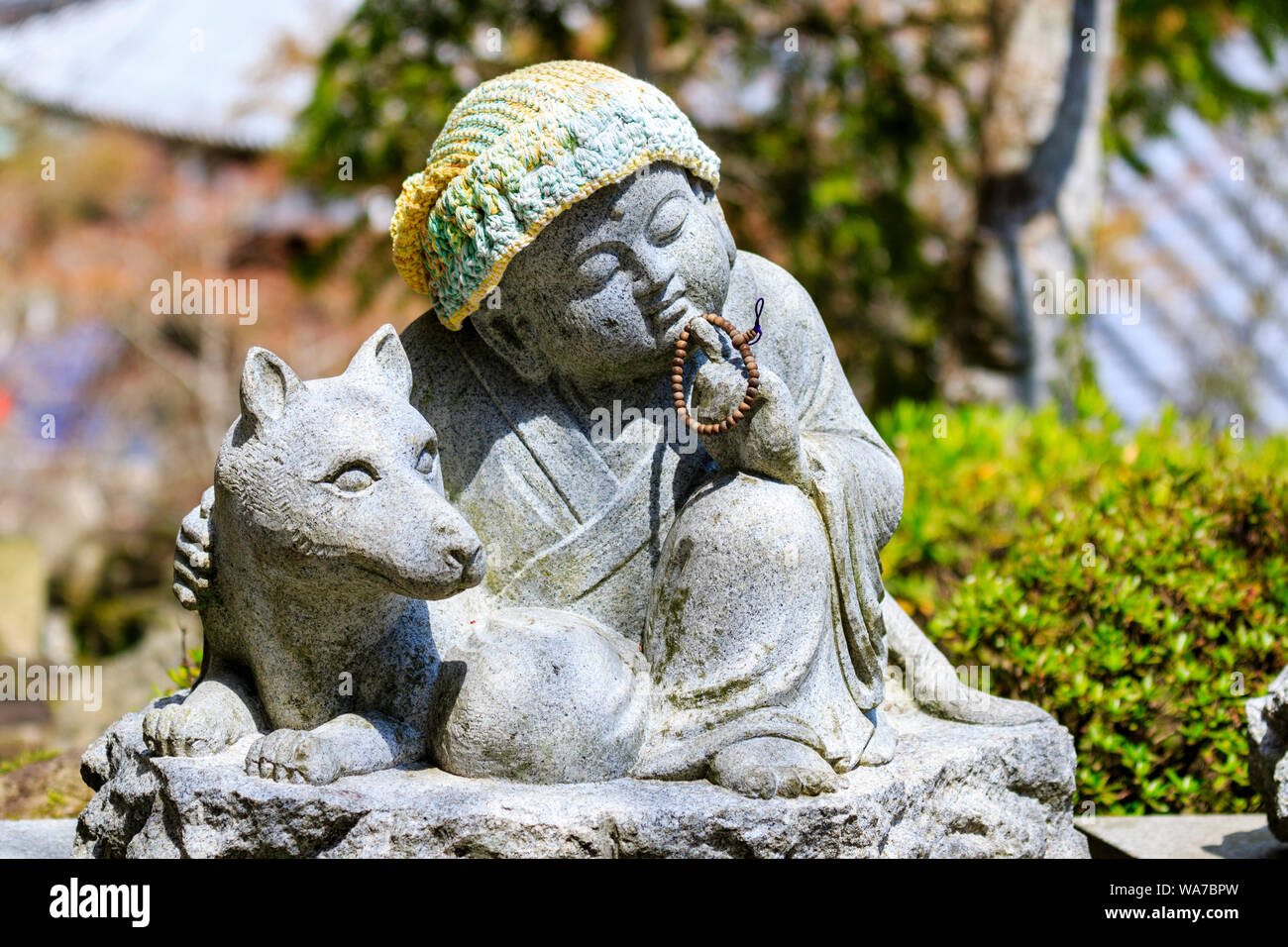 Japan, Miyajima. Daisho-in temple. Small Jizo statue of a sitting Buddhist monk in contemplation, holds a rosary in one hand and arm is around a dog. Stock Photo