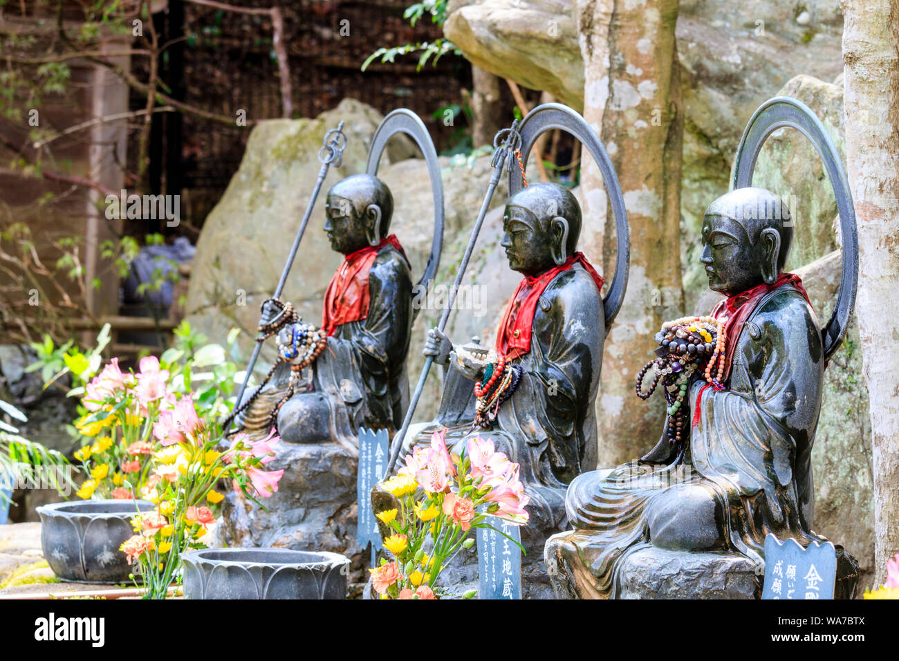 Japan, Miyajima. Daisho-in temple. Line of three Jizo Bosatsu stone statues, two holding scepter, shakujo, and all with red bibs and flowers in front. Stock Photo